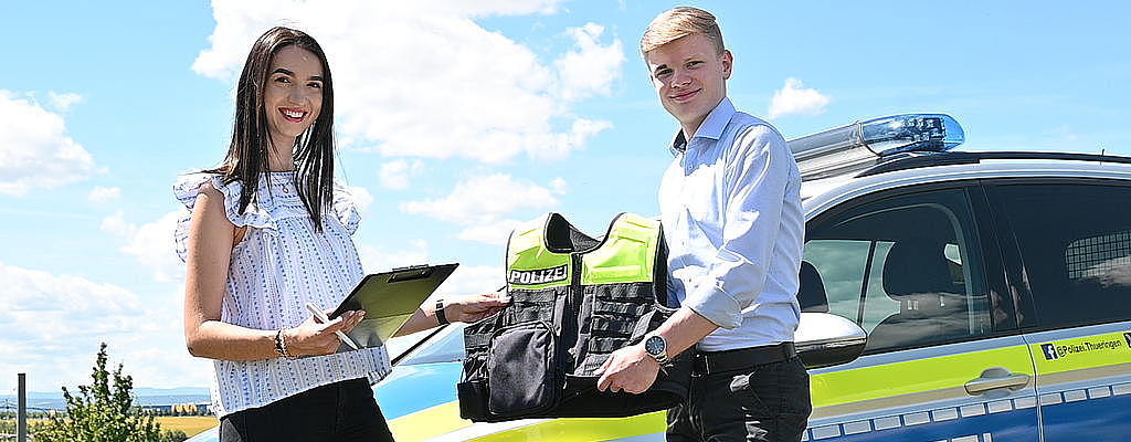 Eine junge Anwärterin und ein junger Anwärter vor einem Polizeiauto